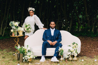 groom sitting on couch while bride stands behind couch around flowers in the forest