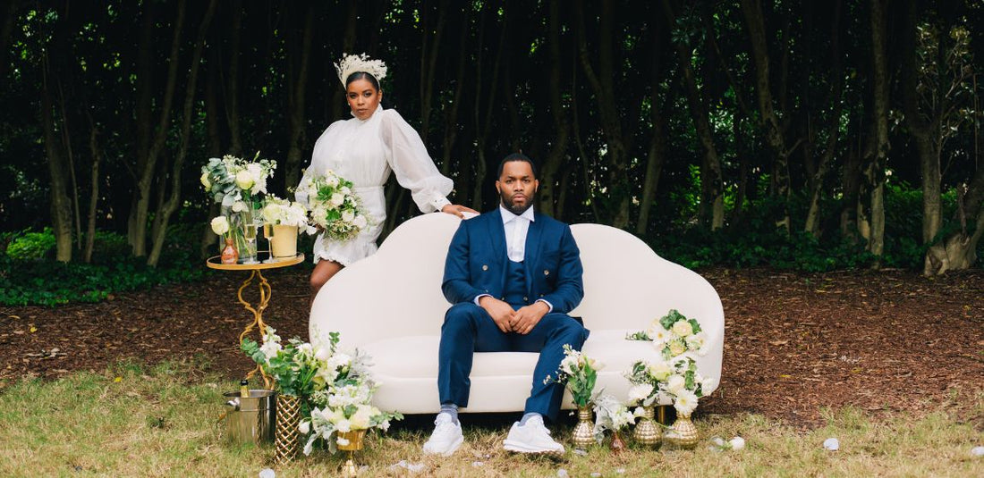 groom sitting on couch while bride stands behind couch around flowers in the forest