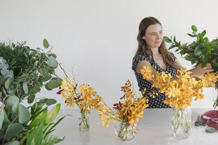 girl arranging greenery and yellow orchids in a vase on a white table