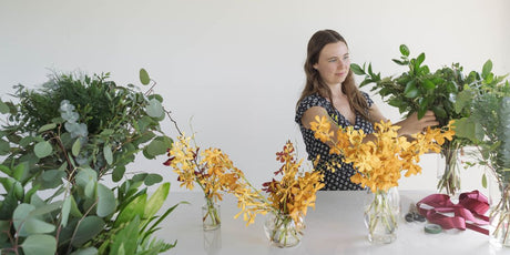 girl arranging greenery and yellow orchids in a vase on a white table